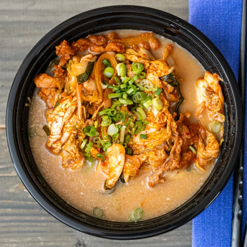 A black bowl of Rai Rai Ramen's kimchi ramen with green onions on a wooden table.